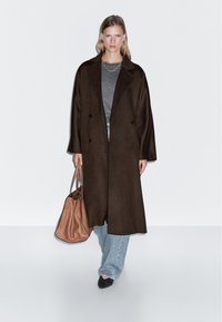 Langer brauner Mantel mit zwei Reihen von Knöpfen, kombiniert mit einem grauen Oberteil und hellen Jeans. Hält eine braune Handtasche; schwarze Schuhe. Schlichte Hintergrund.