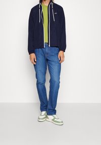 Marinblå vindjacka med dragkedja och vita dragsnören, bärs över en grön t-shirt, tillsammans med blå jeans och sneakers med gröna detaljer.