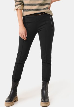 Kvinde iført tætsiddende sorte bukser med lynlåslommer, beige og sort stribet sweater og sorte læderankelstøvler med brune såler.