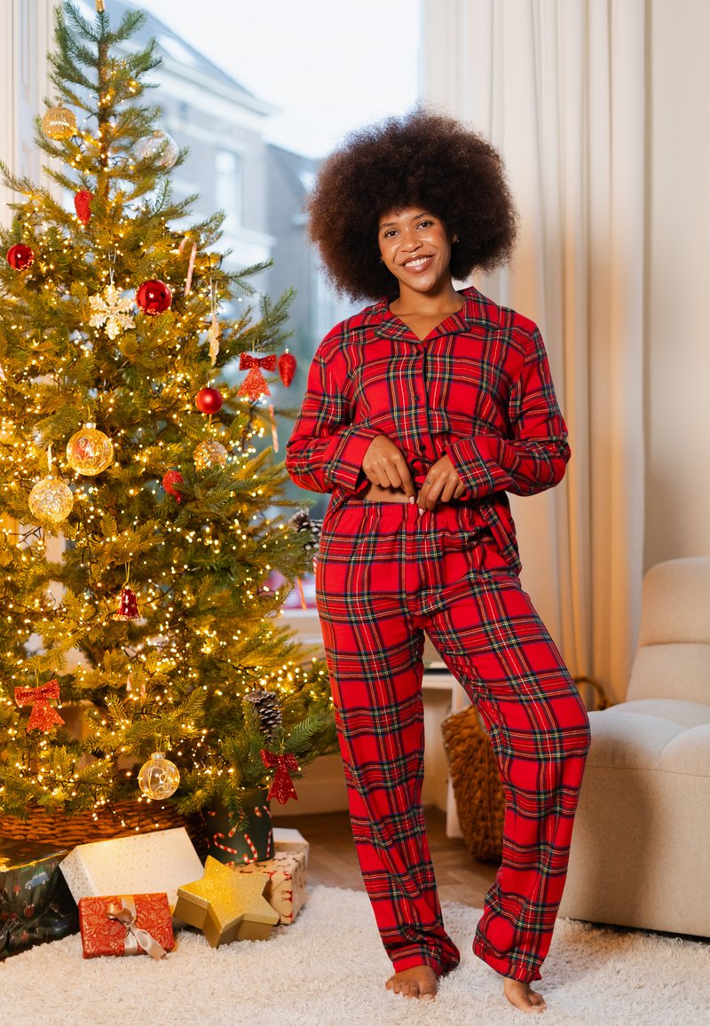 Rood geruit pyjama set bestaande uit een overhemd met knopen en lange mouwen, en bijpassende broek, tentoongesteld in een gezellige, sfeervol ingerichte binnenruimte.