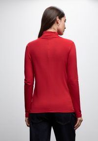 Maglietta rossa a maniche lunghe con colletto alto, realizzata in un tessuto morbido, caratterizzata da una consistenza liscia e una cucitura posteriore dritta. Abbinata a jeans scuri.