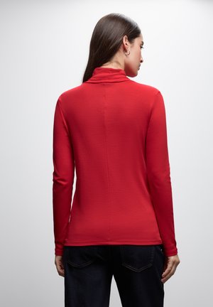 Top rojo de manga larga con cuello alto, hecho de una tela suave, con una textura lisa y una costura trasera recta. Combinado con jeans oscuros.