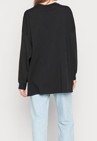 Svart oversized långärmad skjorta med rund halsringning, med lös passform och ribbade ärmslut, som bärs med ljusblå jeans.