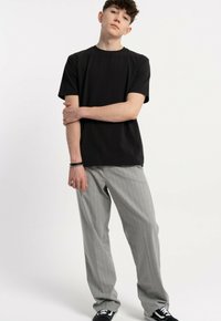 Młody mężczyzna stoi w casualowy sposób, ubrany w czarną koszulkę, szare spodnie w prążki oraz czarne sneakersy z białymi sznurówkami, na jednolitym białym tle.