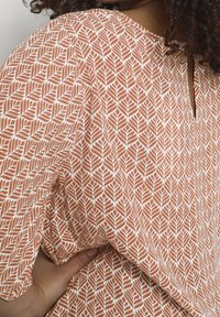 Close-up of a person wearing an orange and white patterned shirt with a small back keyhole opening, hand resting on their hip.