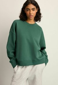 Groene katoenen sweatshirt met een relaxte pasvorm, ronde hals en geribde manchetten, gecombineerd met lichtgrijze joggingsbroek. Zachte textuur en casual ontwerp.