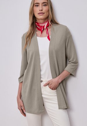 Kvinde iført beige cardigan med åben front, hvid top, hvide bukser og rødt halsklæde med striber, stående mod en lys baggrund.
