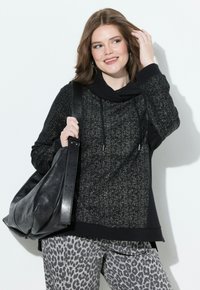 Mujer con cabello hasta los hombros, vistiendo una sudadera negra con manchas y pantalones grises con estampado de leopardo, sosteniendo un gran bolso de cuero negro en su hombro.