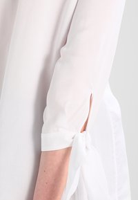 Blouse blanche avec une texture transparente, présentant un détail de manche nouée et une fente au poignet. Tissu doux avec un aspect léger et aérien.