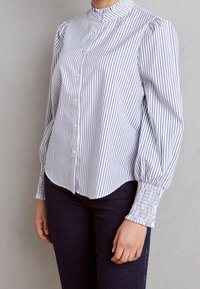 Chemise à rayures blanche et bleu clair, avec un col froncé, des manches ballons et des poignets smockés. Coupe ajustée avec un ourlet arrondi.