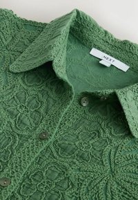 Chemise verte boutonnée avec une broderie en dentelle florale complexe et un col plié, avec une étiquette blanche portant le nom de la marque « NEXT ».
