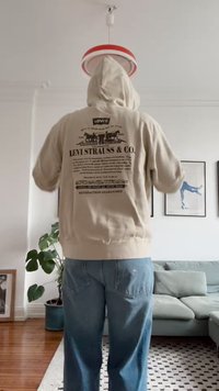 Beige kapucnis pulóver, hátán nyomott szöveggel és grafikákkal. Világoskék farmernadrággal viselve, benti környezetben.