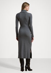 Grå långärmad klänning med hög krage, figurnära silhuett och sidokilar. Modellen bär svarta knähöga stövlar. Slät tygtextur.
