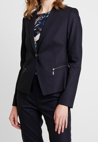 Laivastonsininen bleiseri istuvuudella, jossa on kaksi vetoketjullista taskua ja yksinkertainen nappisulku. Valmistettu sileästä kankaasta, jossa on jämäkät olkapäät.