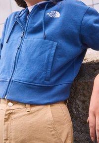 Felpa blu con zip frontale, polsini a costine e logo The North Face in bianco. Indossata con pantaloni di cotone beige con tasche laterali.