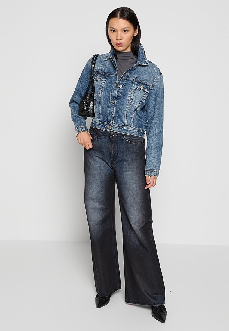 Chaqueta de mezclilla en azul medio con un diseño cropped, combinada con jeans oscuros de pierna ancha y alta cintura. La modelo sostiene un pequeño bolso negro.
