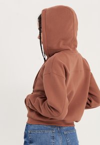 Sudadera con capucha marrón de corte holgado, puños y dobladillo acanalados, combinada con jeans de mezclilla azul de cintura alta. Textura lisa, diseño minimalista.