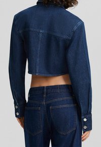 Avkortad mörk denimjacka med spetshals, knäppta manschetter och en ryggsömsöm. Har en ren finish och strukturerad design.