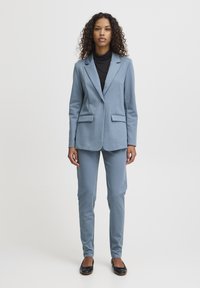 Lichtblauw op maat gemaakt pak met een blazer met één knoop en schuine zakken, gecombineerd met bijpassende slim-fit broek. Zwarte turtleneck eronder.