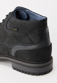 Botín negro con paneles de cuero texturizado y ante, cuello acolchado, forro interior de mezclilla azul y etiqueta con la marca "Lee" en el costado.