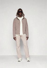 Moški nosi bež kapuco, usklajene hlače, belo majico, bele superge in velike črne sončne očala, stoji ob enotni svetli ozadju.