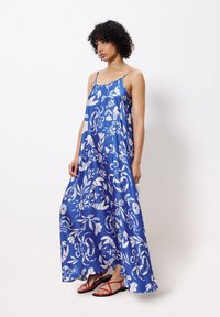 Maxi-jurk van blauwe satijn met witte bloemmotieven, voorzien van dunne verstelbare bandjes en een losse pasvorm, gecombineerd met rode sandaaltjes met bandjes.