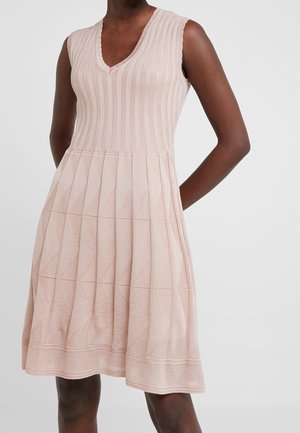 Ærmeløs, knælang lyserød strikket kjole med V-hals og tekstureret geometrisk mønster, båret af en person med mørk hud.