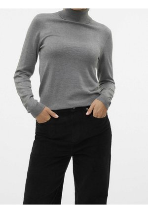 Kvinde iført en grå rullekrave sweater og sorte bukser med hænderne i lommerne, foran en ensfarvet hvid baggrund.