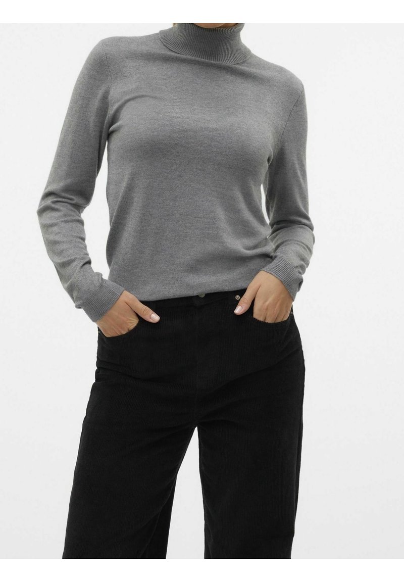 Femme portant un pull à col roulé gris et un pantalon noir, les mains dans les poches, sur fond blanc uni.