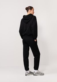 Czarna bluza z kapturem z sznurkiem, w zestawie z dopasowanymi dresami. Posiada elastyczne mankiety i luźny krój. Noszona z srebrnymi butami sportowymi.