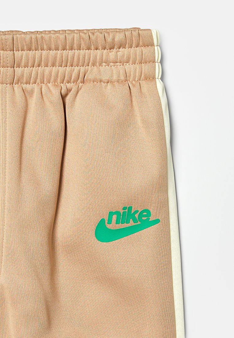 Beżowe spodnie dresowe Nike z elastycznym pasem, białymi paskami po bokach i zielonym logo na udzie, wykonane z teksturowanej tkaniny i w luźnym kroju.
