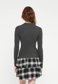 Grå ribbet genser med lange ermer og en tettsittende design, kombinert med et svart og hvitt rutete skjørt med volanger. Ren hvit bakgrunn.