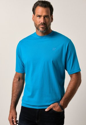 Camiseta de manga corta turquesa en algodón suave, con un sutil logo en relieve "JP" en el pecho, combinada con jeans oscuros.