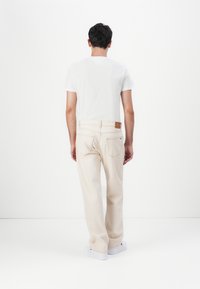 Pantalones de pierna recta color crema, hechos de algodón, con dos bolsillos traseros y un acento de cinturón de cuero marrón. Combinados con una camisa blanca.