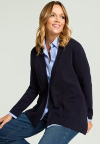 Marineblauer Strickcardigan mit Knopfverschluss, gerippten Bündchen und zwei Fronttaschen, getragen über einem hellblauen Hemd mit Kragen.