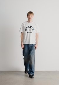 Valge puuvillane T-särk tumesinise trükiga, millel on kiri "CALVIN KLEIN", koos helesiniste teksadega. Mudel kannab musti kingi, seistes heledal hallil põrandal.