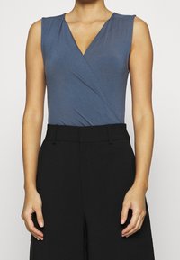 Modrý bezramínkový body s hlubokým V-výstřihem a zavinovacím designem, spojený s vysokými černými kalhotami, které vystavují hladkou, přiléhavou texturu.