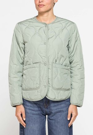Donna che indossa una giacca trapuntata verde chiaro con bottoni a pressione e due tasche frontali, abbinata a jeans blu, in piedi davanti a uno sfondo chiaro.