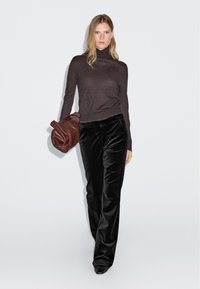 Brun polotröja, svarta vida byxor i sammet och vinröd handväska. Släta texturer, ledig passform, stående pose mot enfärgad bakgrund.