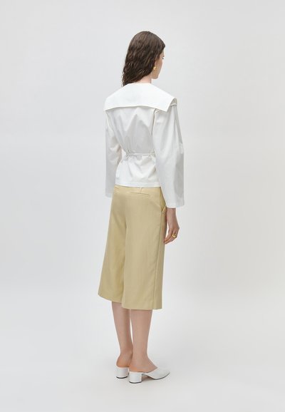 Kobieta o kręconych brązowych włosach nosi biały top z długim rękawem i szerokim kołnierzem, beżowe spodnie do kolan, białe mules na obcasie oraz złote kolczyki.