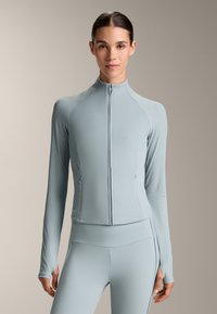 Svijetloplava atletska jakna s patentnim zatvaračem, rebrastom teksturom, dugim rukavima i prilagođenim dizajnom, sadrži zatvaranje na patentni zatvarač i bočne džepove.