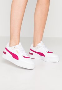 Personne portant des baskets blanches Puma avec des rayures latérales rose vif, se tenant sur un sol clair et uni devant un fond neutre.