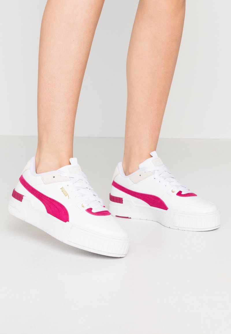 Personne portant des baskets blanches Puma avec des rayures latérales rose vif, se tenant sur un sol clair et uni devant un fond neutre.