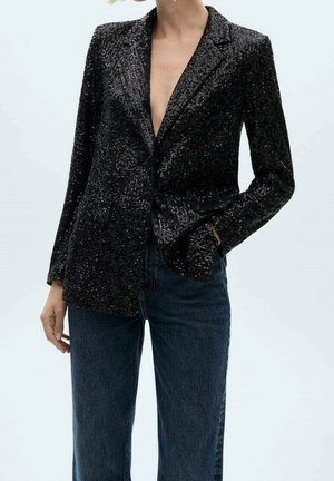 Frau trägt einen schwarzen Paillettenblazer mit tiefem V-Ausschnitt und dunkelblauer Jeans, posiert mit einer Hand in der Tasche.