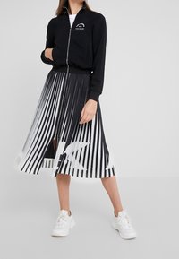 Schwarze Reißverschlussjacke mit weißem Logo, kombiniert mit einem schwarz-weißen plissierten Rock mit Streifenmuster. Weiße Turnschuhe vervollständigen das Outfit.