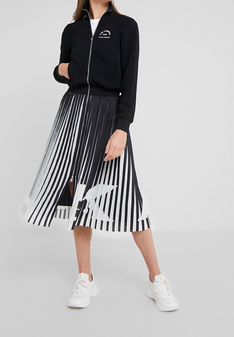 Schwarze Reißverschlussjacke mit weißem Logo, kombiniert mit einem schwarz-weißen plissierten Rock mit Streifenmuster. Weiße Turnschuhe vervollständigen das Outfit.