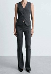 Op maat gemaakte grijze vest met een V-hals en drie knopen, gecombineerd met strakke grijze broeken met zijnaad en puntige zwarte hakken.