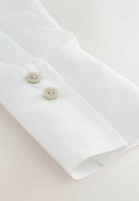 Bílý manžetový límec košile s dvěma světle zbarvenými knoflíky, hladkou texturou látky a čistým šitím podél okrajů.