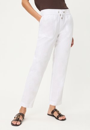 Persona che indossa pantaloni bianchi con coulisse e tasche frontali e sandali neri a punta aperta, in piedi davanti a uno sfondo chiaro e uniforme.