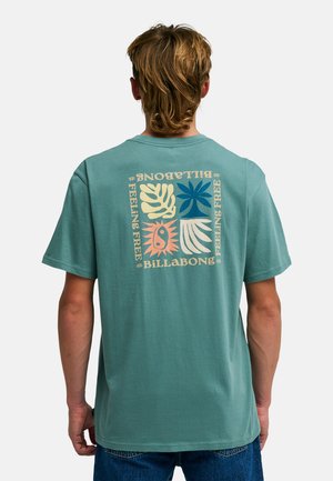 Uomo con capelli biondi che indossa una maglietta verde acqua a maniche corte con disegni astratti colorati di foglie e sole e la scritta "Billabong" sulla schiena.